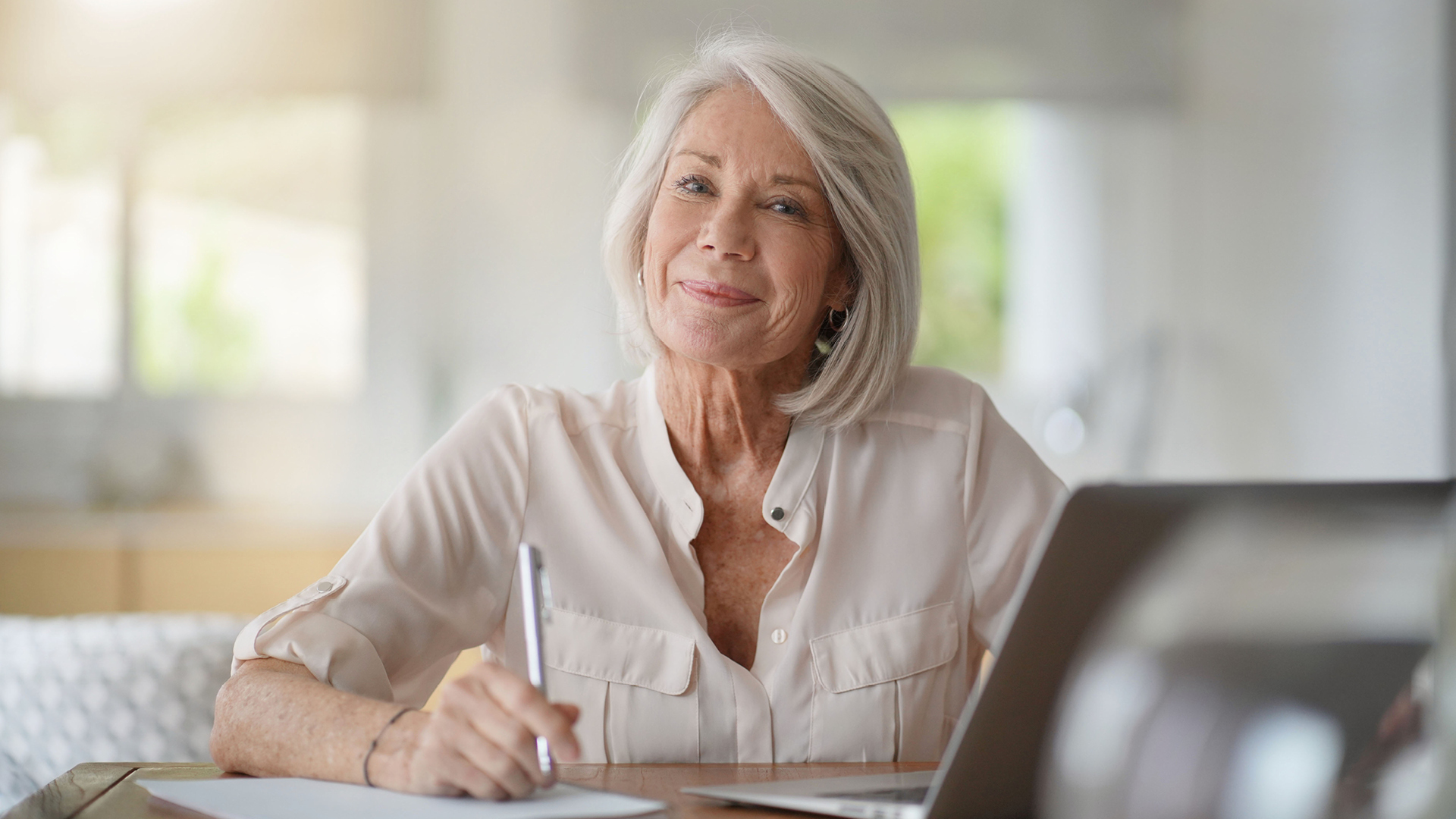 mature woman writing on paper at a desk looking at camera